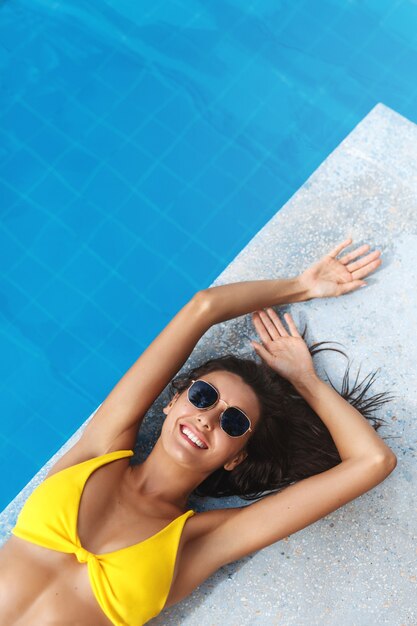 Top view of beautiful brunette woman with golden summer tan, lying near swimming pool and smiling in sunglasses and bikini.