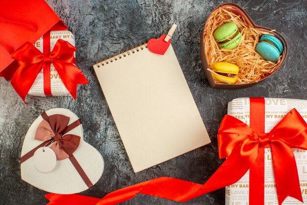 Top view of beautiful gift boxes tied with red ribbon and delicious macrons in heart shaped box spiral notebook on icy dark background