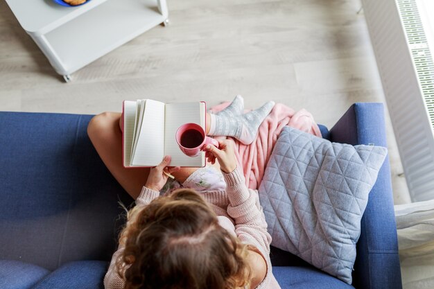 Top view of beautiful young woman holding cup  of tea while relaxing on couch at home. Warm cozy morning time.