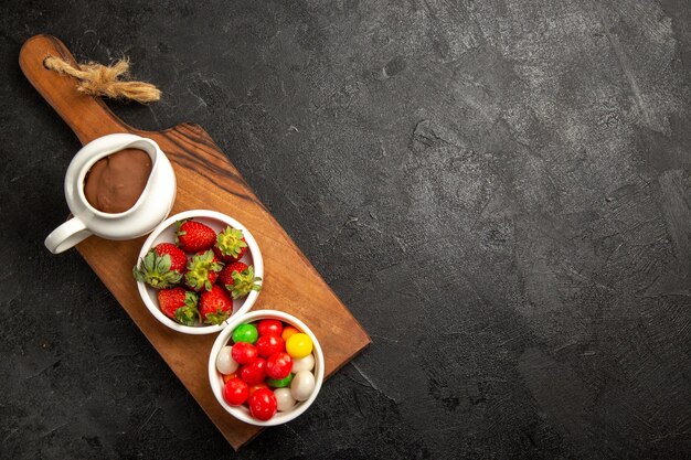 어두운 테이블의 왼쪽에 있는 나무 커팅 보드에 있는 달콤한 초콜릿 크림과 딸기의 상위 뷰 베리 그릇