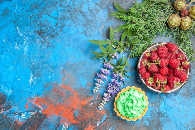 Top view of berry bowl, small tart and tree branches on blue surface