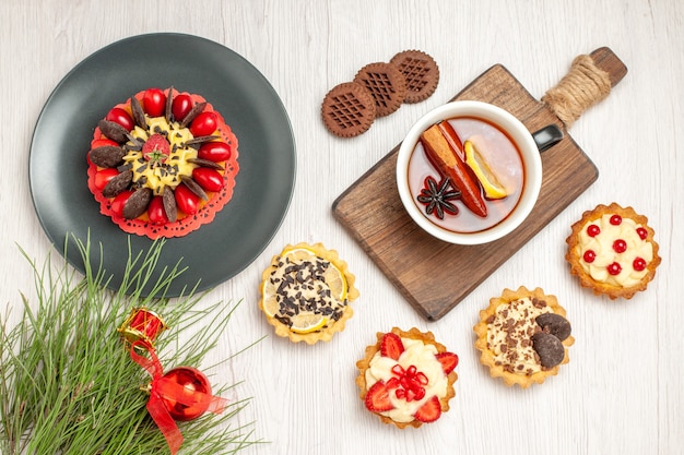 Top view berry cake in a grey plate tarts a cup of lemon cinnamon tea on the chopping board cookies and the pine tree leaves on the white wooden ground