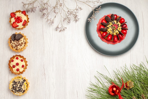 Top view berry cake in a grey plate at the top right and from top to the bottom different tarts at the left and the pine tree leaves at the bottom right of the white wooden ground with copy space