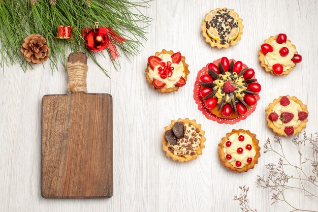 Top view berry cake rounded with tarts pine tree leaves with christmas toys and a chopping board on the white wooden ground