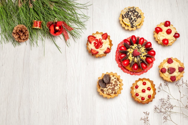 Top view berry cake rounded with tarts and pine tree leaves with christmas toys on the white wooden ground