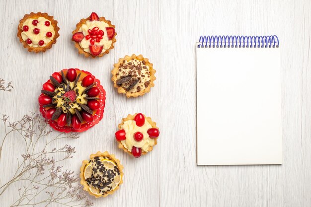 Top view berry cake surrounded berry and chocolate tarts at the left and a notebook at the right side of the white wooden ground