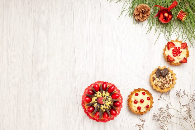 Top view berry cake tarts and the pine tree leaves with christmas toys at the right side of the white wooden table