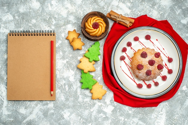 Top view berry cake on white oval plate red shawl notebook red pencil cookies on grey surface
