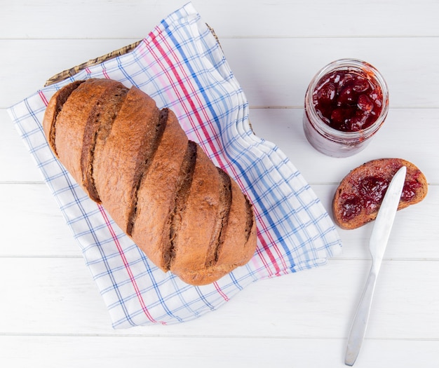 Top view of black bread on plaid cloth with jam smeared on piece of rye bread and knife on wooden table