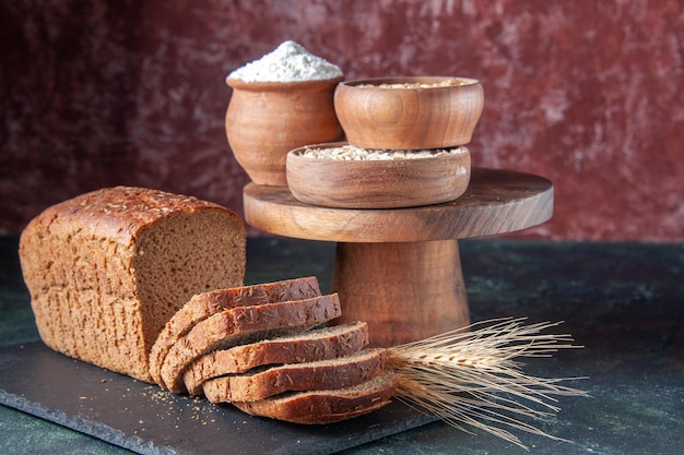 Top view of black bread slices on dark color tray flour oatmeal buckwheat on wooden board on mixed color distressed background