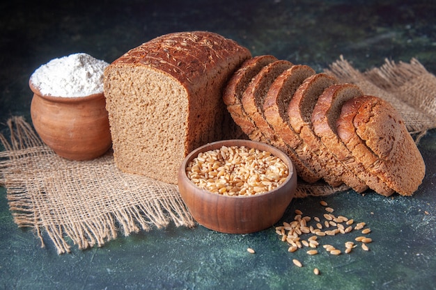 Top view of black bread slices flour in a bowl and wheat on nude color towel on maroon color background