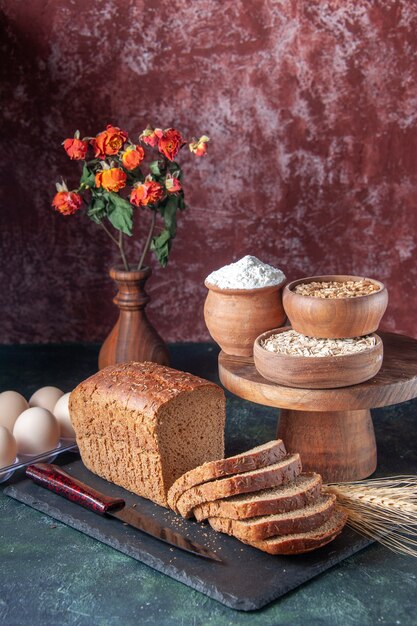 Top view of black bread slices knife on dark color tray flour oatmeal buckwheat on wooden board eggs on mixed color distressed background