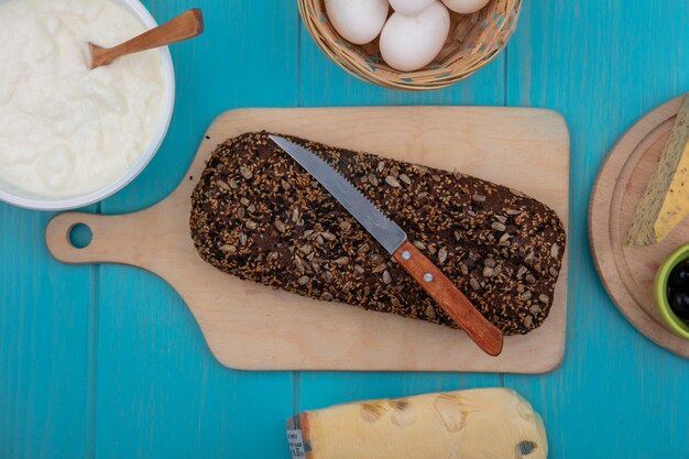 Top view black bread with knife on cutting board and chicken eggs with yogurt in bowl on turquoise background