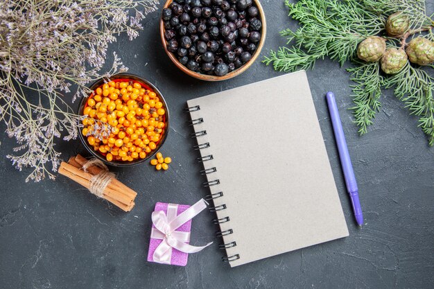 Top view black currant sea buckthorn in bowls small gift notepad pen cinnamon sticks on dark surface