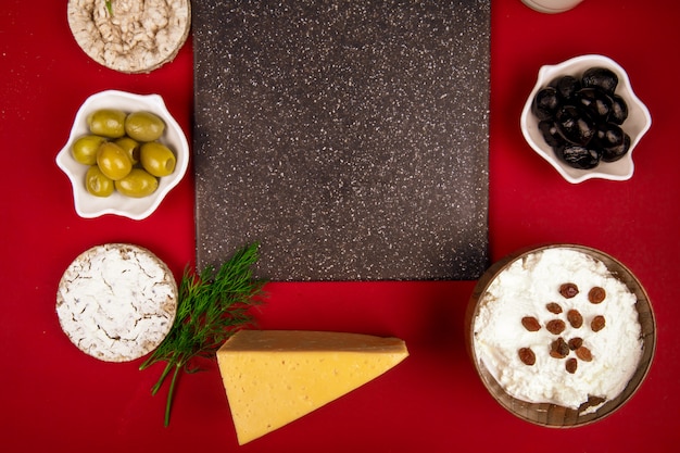 Вид сверху черная разделочная доска и маринованные оливки стакан молока творога в миску козьего и голландского сыра, расположенных вокруг на красный