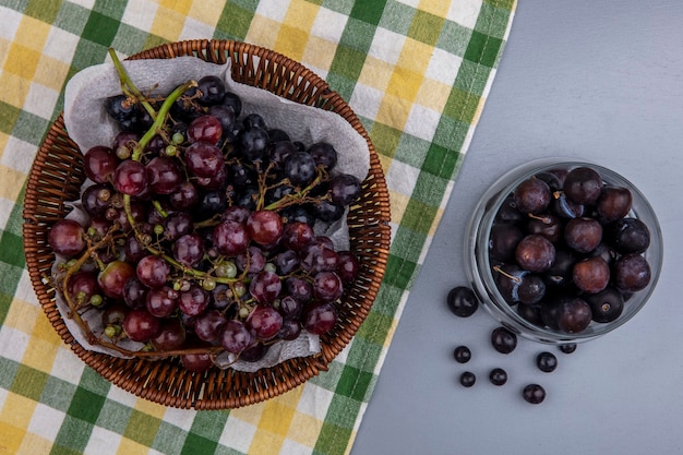 Вид сверху черного винограда в корзине на клетчатой ткани и виноградных ягод в миске на сером фоне
