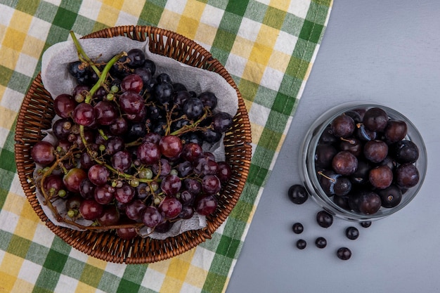 Top view of black grape in basket on plaid cloth and grape berries in bowl on gray background