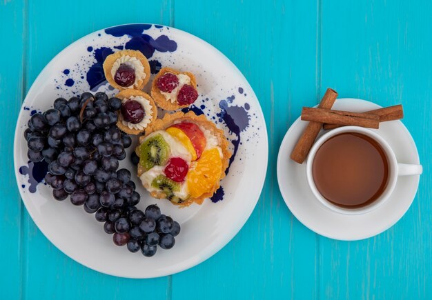 Top view of black grapes on a plate with a cup of tea and cinnamon sticks on a blue wooden background