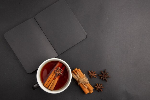 Top view black notebook cup of tea flavored by cinnamon and anise cinnamon sticks tied with rope on dark table