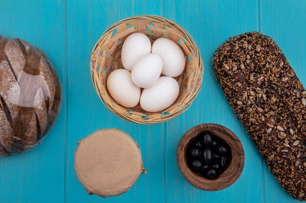 Top view black olives in a bowl with chicken eggs in a basket and black bread on a turquoise background