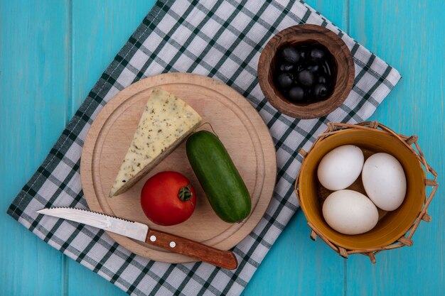 Top view black olives with cheese  cucumber  tomato and chicken eggs in a checkered towel  on a turquoise background