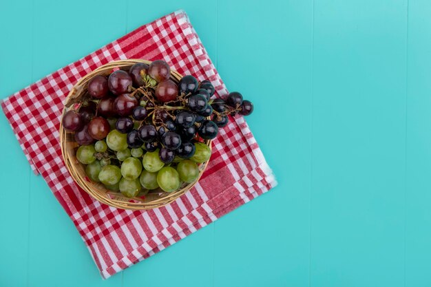 Top view of black red and white grapes in basket on plaid cloth on blue background with copy space