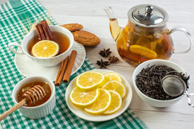 Top view of black tea in a white cup and a glass pot with cinammon limes and lemon next to delicious cookies honey on green stripped towel on white table