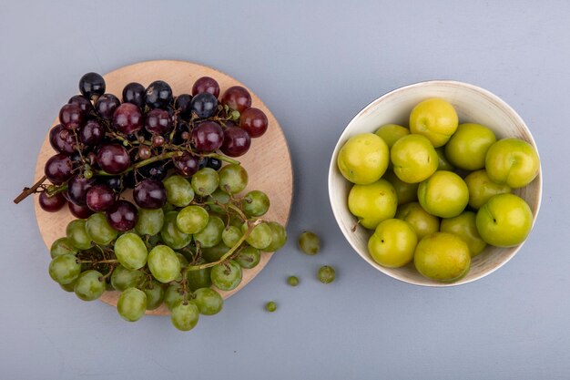 Top view of black and white grapes on cutting board and bowl of plums on gray background