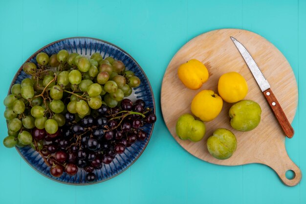 Top view of black and white grapes in plate and green pluots apricots with knife on cutting board on blue background