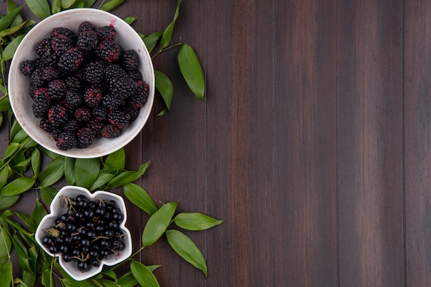 Top view of blackberry in a bowl with black currant and leaf branches on a wooden surface