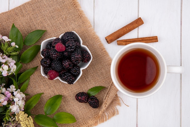 Top view of blackberry in a bowl with flowers and a cup of tea with cinnamon on a white surface