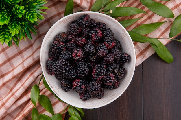 Top view of blackberry in a bowl with leaf branches on a checkered brown towel