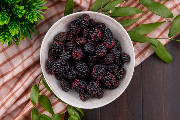 Top view of blackberry in a bowl with leaf branches on a checkered brown towel