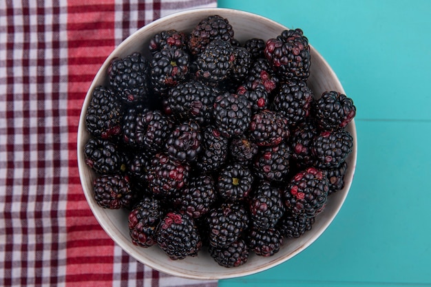 Top view of blackberry in a bowl with a red checkered towel on a turquoise surface