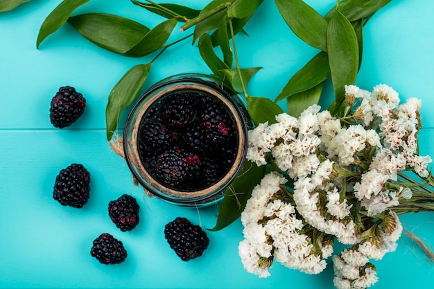 Top view of blackberry in a jar with a branch of leaves and white flowers on a light blue surface