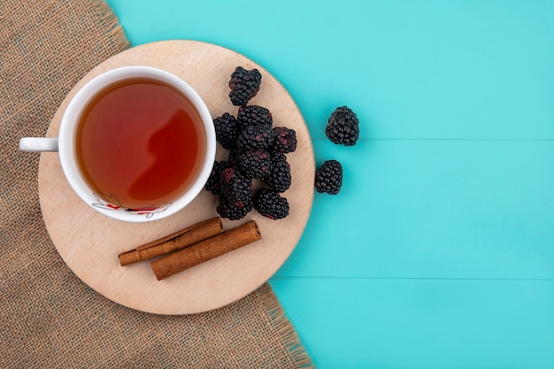 Top view of blackberry with a cup of tea and cinnamon on a stand on a turquoise surface