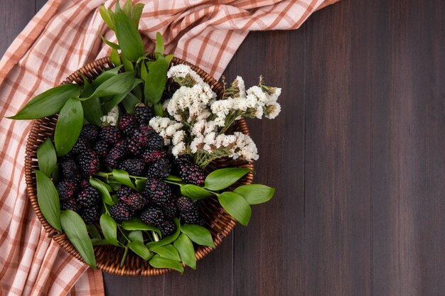 Top view of blackberry with white flowers in a basket with a checkered cinnamon towel on a wooden surface