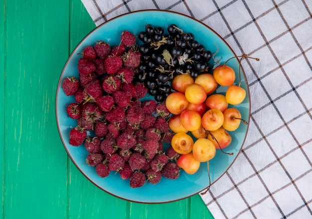 Top view of blackcurrant with raspberries and white cherries on a blue plate with a checkered towel on a green surface