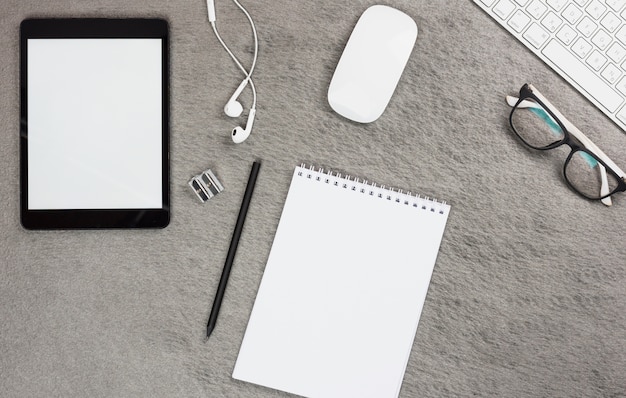 Top view of blank digital tablet; earphone; mouse; keyboard and eyeglasses with spiral notepad