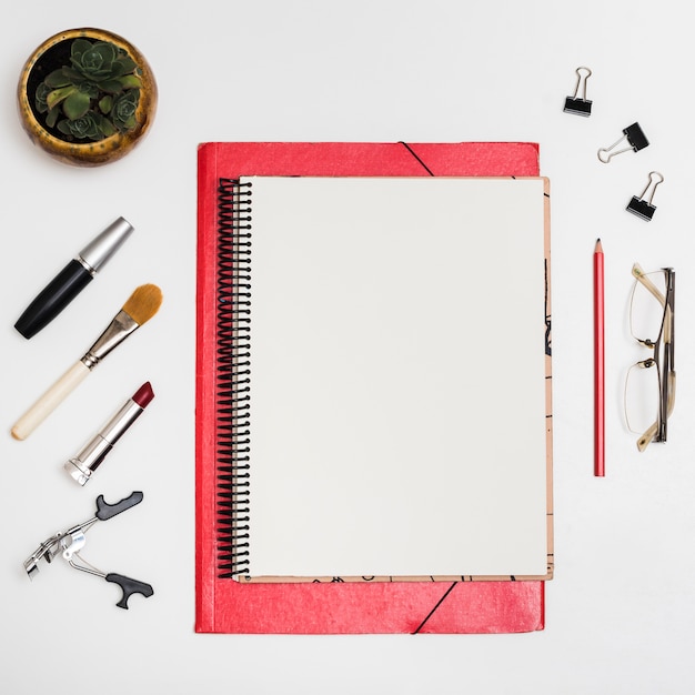 Top view of blank notebook with cosmetic products; paper clips; pencil; eyeglass over white desk