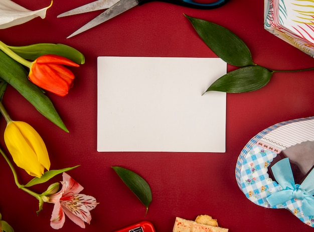 Вид сверху чистого листа поздравительной открытки и тюльпана с цветами альстромерии с подарочной коробкой в форме сердца на красном столе