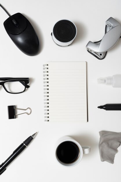 Top view of blank spiral notepad with office stationery; coffee cup; eyeglass over white desk