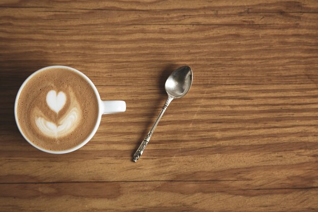 Top view on blank white cup with cappuccino with silver spoon on thick brutal wooden table in cafe shop. Foam with heart shape. Focus on top cup