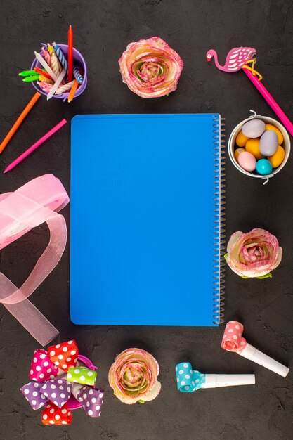 A top view blue copybook with flowers candies pencils all around on the dark desk color candy photo book