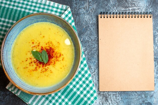 Top view of a blue pot with tasty soup served with mint on half folded green stripped towel next to spiral notebook on blue background