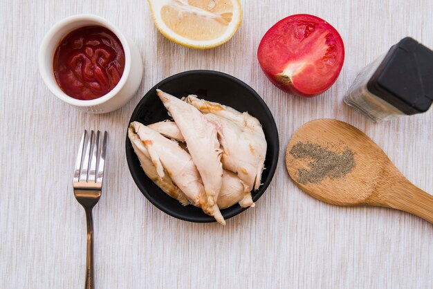 Top view of boiled chicken in black plate with black pepper powder; tomato; lemon; fork and tomato sauce over wooden desk