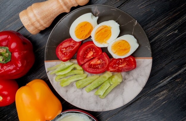 Top view of boiled eggs on a plate with tomato slices and colorful bell peppers on wooden background