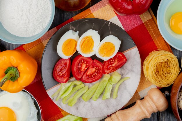 Top view of boiled eggs on a plate with tomatoes and slices of green bell peppers on a checked tablecloth on a wooden background