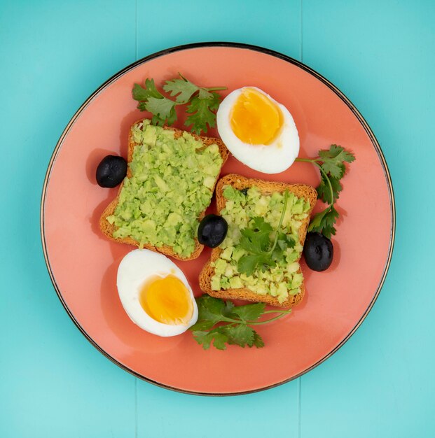 Top view of boiled eggs with a toasted bread woth avocado pulps with olives onn orange plate on blue