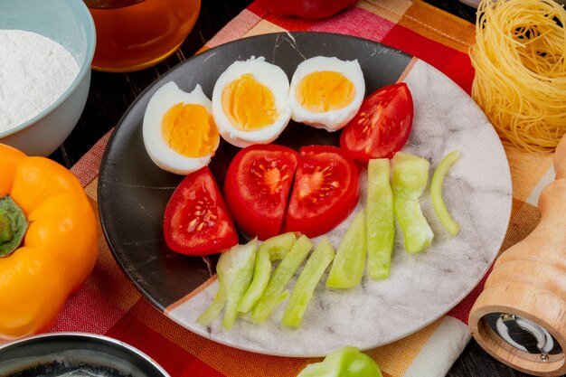 Top view of boiled halved eggs on a plate with slices of tomatoes and bell peppers on a checked tablecloth on a wooden background